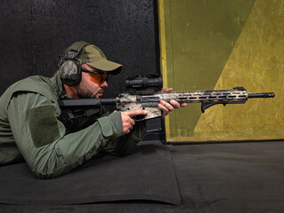 Fototapeta premium A focused male shooter in tactical clothing, safety glasses, and earmuffs aims a modern camouflage rifle from a prone position.