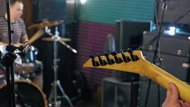 Close-up of the neck of an electric guitar with a musician playing drums in the background in a bright studio. View from behind the guitarist. The studio has colored soundproofing panels