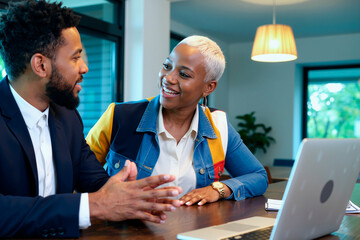 Fototapeta na wymiar Young adult Black man and young adult Black woman sitting at table discussing business strategy, smiling and making eye contact, open laptop and documents visible in modern office setting