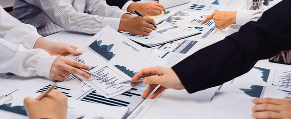 A business meeting scene showing a diverse team engaging with data charts and reports on the table, alongside laptops and digital screens in a modern office setting. SACTR