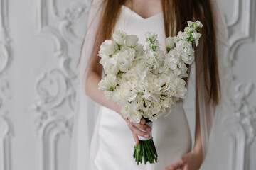 Bride in a white satin wedding dress holding a lush, vertical white bouquet of stock flowers and eustoma carnations against a background of classic white stucco wall paneling