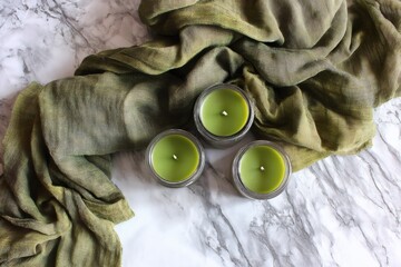 Three green scented candles in glass jars with a draped fabric on white marble, top view, calm atmosphere