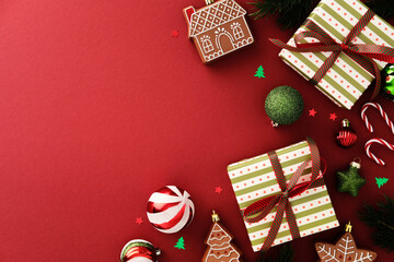 Christmas flat lay with gifts, gingerbread cookies, ornaments, candy canes, and pine branches on red background. Warm festive composition for seasonal greeting or holiday design.