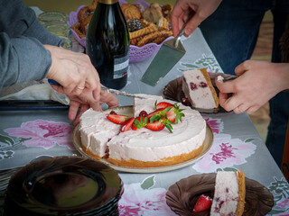 Friends come together in a warm home, enjoying a delicious dessert. They cut into a creamy cake topped with fresh strawberries, surrounded by treats and drinks.