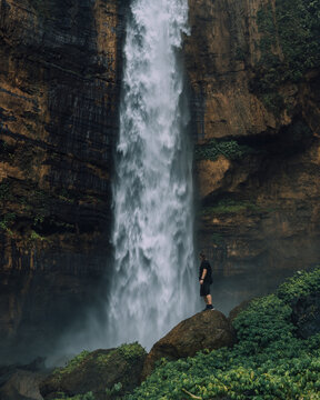 View of a lone figure silhouetted against the misty cascade of a powerful waterfall amidst rugged cliffs and lush greenery, Kapas Biru Waterfall, Jawa Timur, Indonesia.