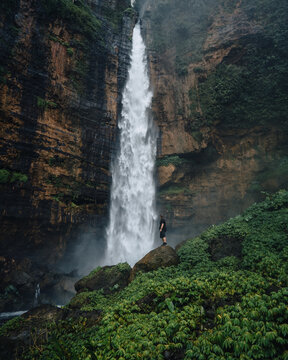 View of a stunning waterfall cascading down rugged cliffs, a solitary figure stands on a mossy boulder, mist rising in Kapas Biru, Jawa Timur, Indonesia.