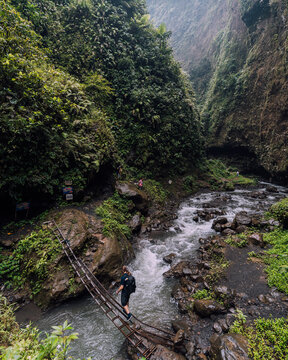 View of a lone figure carefully crossing a rickety bamboo bridge over the rushing, rocky river in a verdant, tropical gorge, Tumpak Sewu, Jawa Timur, Indonesia.