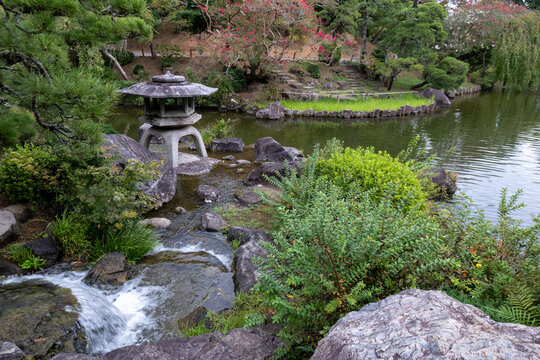 View of a serene Japanese garden with a stone lantern, flowing water, and vibrant greenery reflecting on the tranquil pond's surface, Narita, Chiba, Japan.