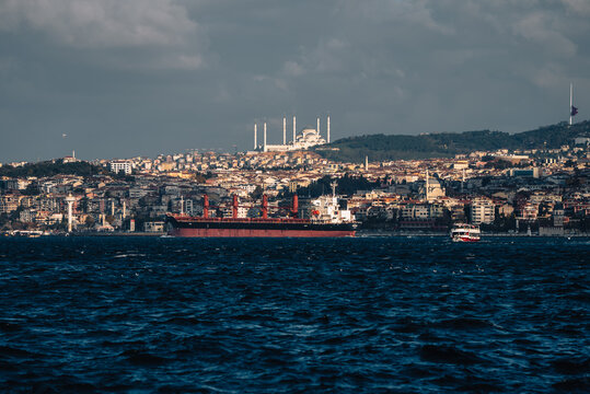 View of a vast, dark sea reflecting the cloudy sky, with a distant cityscape featuring buildings, a mosque, and a large ship sailing near the shore, Istanbul, &Auml;&deg;stanbul, T&Atilde;&frac14;rkiye.