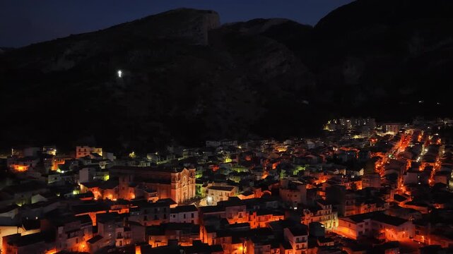 Aerial drone night view of Collesano, a historic Sicilian mountain town at dusk with Basilica San Pietro church and illuminated stone streets in the Madonie Mountains, Sicily, Italy