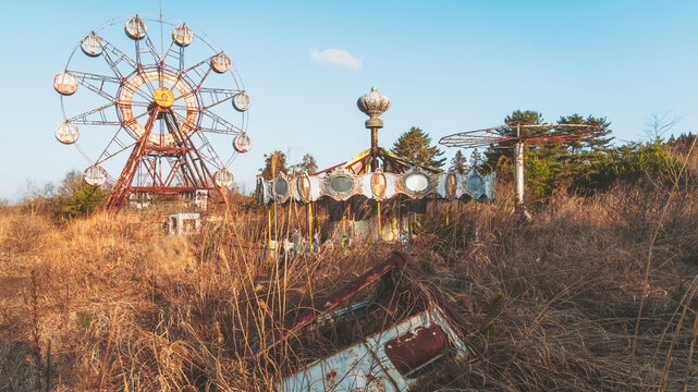 View of a rusted Ferris wheel and decaying carousel stand amidst a field of golden, withered grass under a pale blue sky, Osaki, Miyagi, Japan.
