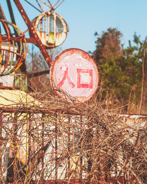View of a dilapidated Ferris wheel looms behind a weathered Japanese sign amidst tangled vines, evoking a sense of forgotten joy, Osaki, Miyagi, Japan.