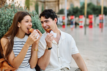 Happy couple  eating a pastry croissant, smiling and looking away on a city street