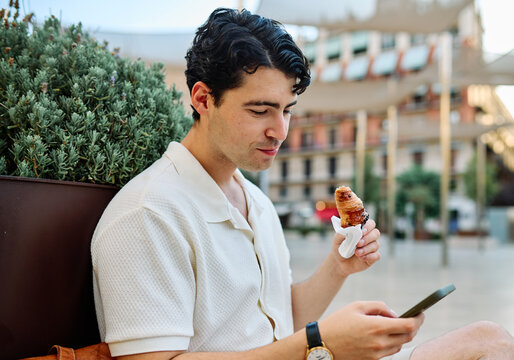 Happy man eating a pastry croissant, smiling and using a mobile phone on a city street