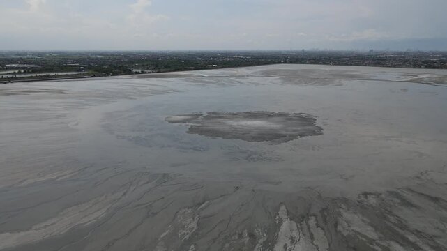Aerial view of the textured mudflow landscape, contrasting with the distant cityscape under a hazy sky, Sidoarjo, Jawa Timur, Indonesia.