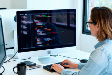 Caucasian young adult woman working at computer monitor, typing code on keyboard, sitting at desk in modern office, focusing on software development, screen displaying programming interface