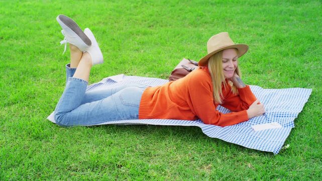 Happy caucasian woman in hat lying on blanket in park, smiling while using smartphone for texting and browsing social media during beautiful sunny day in early autumn