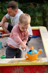 Boy, playing in the sandbox with his dad, fills a watering can with water