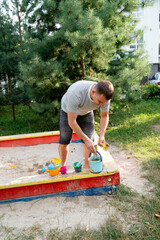 Father collecting toys after playing with children in sandpit at a local park on a sunny afternoon