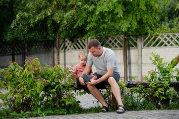 Father and his young child sit on a bench surrounded by greenery. The father wipes his son's feet
