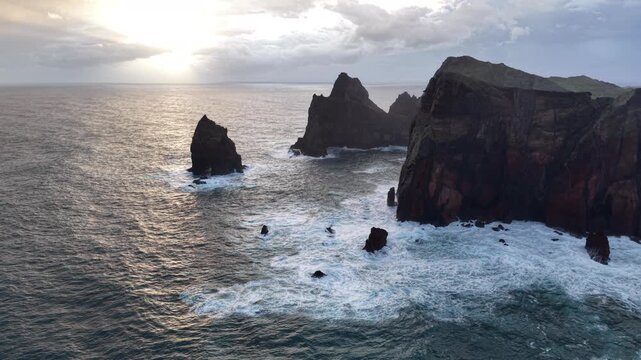 Aerial view of dark cliffs meeting the foamy ocean waves under a cloudy sky, creating a dramatic scene, Canical, Madeira, Portugal.