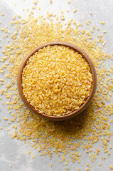 Bowl filled with golden bulgur wheat surrounded by scattered grains on a light surface, showcasing the texture and natural color of this nutritious ingredient