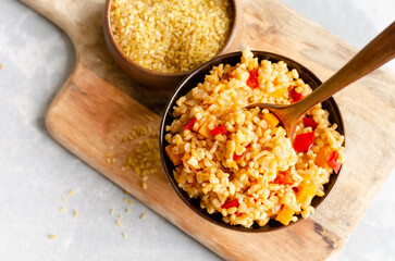 Colorful bowl of cooked bulgur wheat with diced bell peppers, served on a wooden cutting board, accompanied by a small bowl of uncooked bulgur, showcasing healthy grains and vibrant ingredients