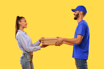 Arabic Courier Guy Giving Pizza Boxes To Female Customer Standing Over Yellow Studio Background....