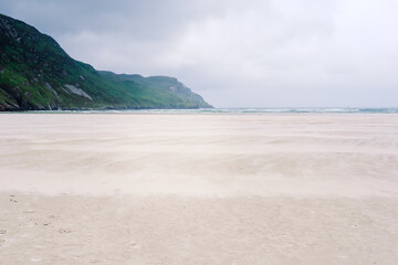 Wind sweeps sand across a pristine beach in Ireland under a moody sky with lush green hills in the background