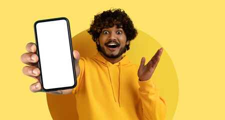 Obraz premium A young man with curly hair is enthusiastically displaying a smartphone with a blank screen. He stands against a vibrant yellow background, conveying joy and excitement.