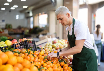 Man shop assistant puts ripe mandarin vegetable in box on display case, arranges assortment in vegetable shop. Worker puts vegetables in pile, in pyramid. Employee makes attractive display case .