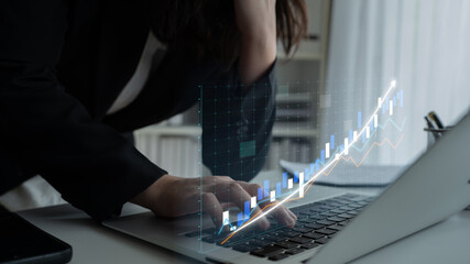 A woman in professional attire focuses on stock market data displayed on her laptop. Digital graphs...