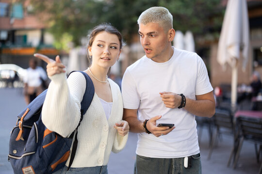 Man holds a mobile phone in hands and shows a map of the city to a female tourist. Woman with a backpack travels to European cities and asks a guy for directions