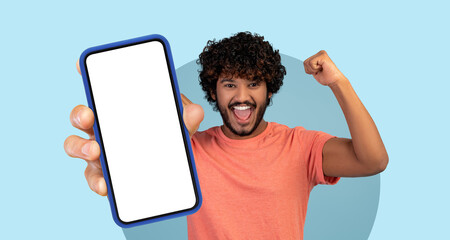 Young man with curly hair smiles broadly while holding a smartphone. He appears joyful and...