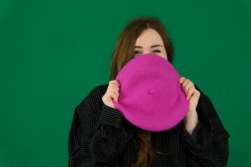 Waist up portrait of a young European woman hiding her nose and mouth under a beret with only her eyes smiling on a green chroma key background with copy space.