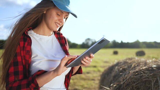 Woman in field holding tablet computer. Girl works on tablet in field. Woman works in a field with haystacks of grass. A woman in a field is lifestyle holding a tablet.