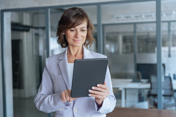 Obraz premium Latin Hispanic mature adult professional business woman looking at online trade app. European businesswoman CEO holding digital tablet using fintech tab application standing at workplace in office.