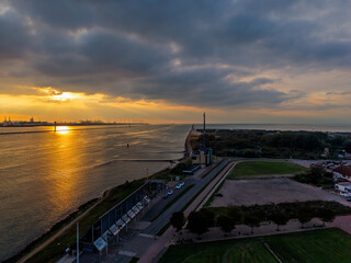 Aerial sunset view of canal and coastline golden light reflects on water as industrial cranes contrast with green fields, road, and rural buildings in a layered twilight landscape.