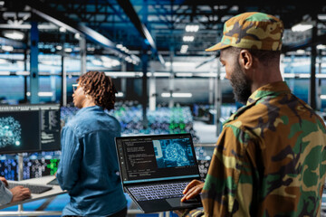 African american military staff uses AI tools for defense systems, working with IT developer in server room. Drone operator in military data center doing live drone monitoring on laptop.