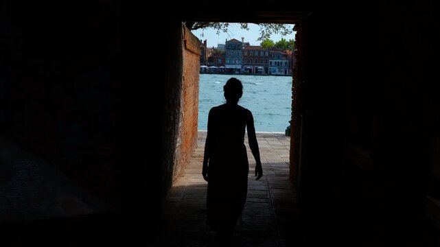 Woman walking towards light in venice. Elegant silhouetted woman walking through historic venetian portico, bathed in warm sunlight with scenic canal backdrop highlighting architectural beauty