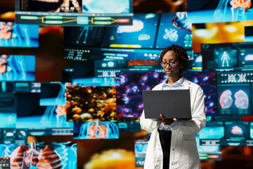 Doctor in lab coat monitoring medical research data and 3D anatomy models dashboards with laptop software. Clinician using notebook to project visual medical reports on multimedia wall