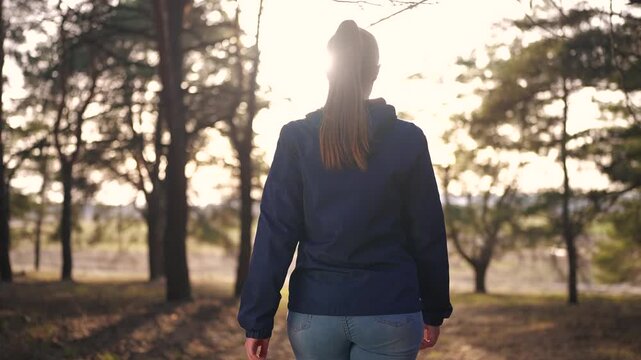 girl tourist walks through the forest. tourist trip concept. a young happy girl in a blue jacket walks through the spring forest with back view, and the tourist glare of the sun lifestyle.
