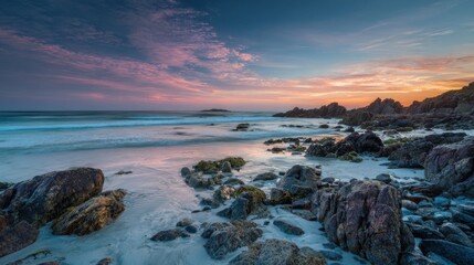 Obraz premium Soft waves gently wash over a rocky beach as the sun sets creating a beautiful blend of colors in the sky. The scene captures the tranquility of nature at dusk.