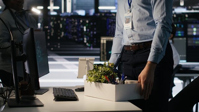 Upset data center worker packing personal items at workstation after being replaced by AI. Programmer removing belongings from server hub workplace desk, fired by artificial intelligence, camera A