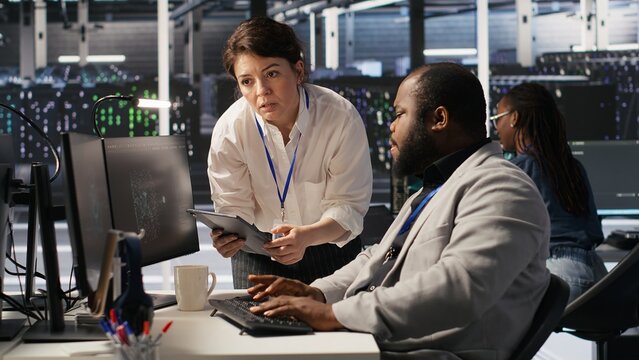 Server room manager supervise technician examining deep learning algorithms. Data center team leader and African american employee review performance dashboards of AI equipment rigs, camera A