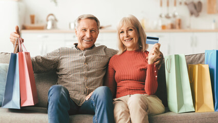 Obraz premium Successful Shopping. Happy Mature Couple Showing Credit Card Sitting With Colorful Paper Shopper Bags On Sofa, Smiling To Camera Posing At Home. Easy Payments And Bank Service Advertisement