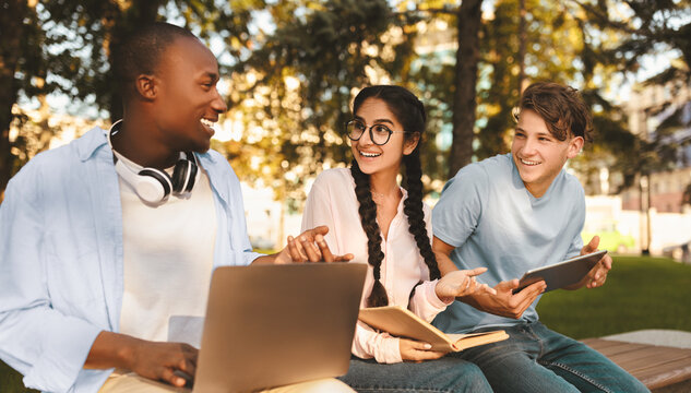 Students lifestyle. Three university friends learning together, using laptops and reading book, sitting on bench in campus park outdoor. Diverse high-school students preapring for exams