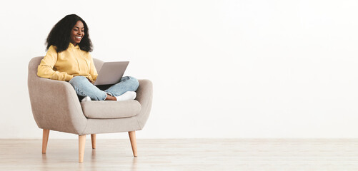 Positive young afro-american woman freelancer working online from home, sitting in comfortable arm chair, typing on laptop keyboard and smiling, chatting with client, white wall background, copy space