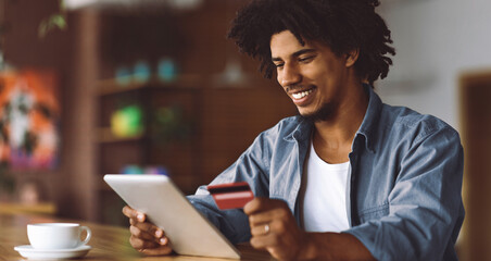 Online Shopping. Young Black Guy Using Digital Tablet And Credit Card While Relaxing In Cafe, Smiling African American Man Drinking Coffee And Purchasing On Internet, Enjoying E-Commerce, Copy Space