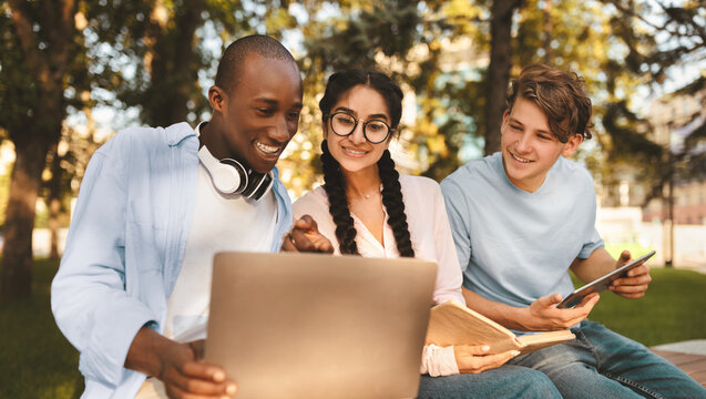 Excited international students studying in park, enjoying the results of their job, looking at laptop screen and smiling, sitting together on bench in campus